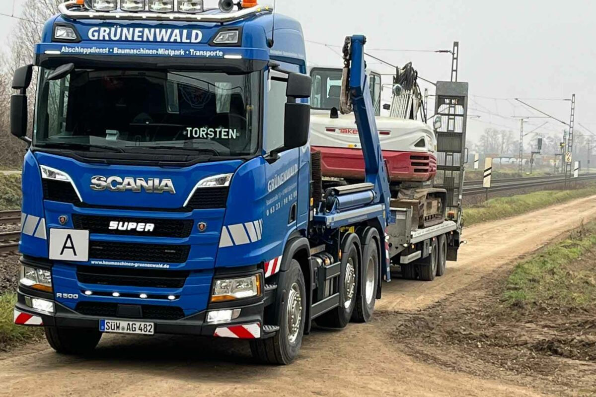 Ein blauer LKW mit der Aufschrift „Grünenwald“ transportiert Baumaschinen auf einem Feldweg in der Nähe von Bahngleisen und bietet zuverlässige Fahrzeugtransporte an.