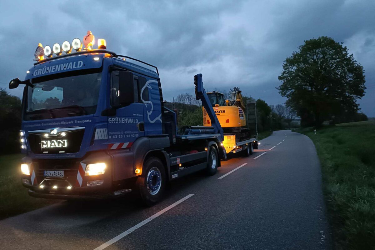 Ein Fahrzeugtransport-LKW transportiert in der Abenddämmerung einen Bagger auf einer Landstraße. Am Himmel hängen dunkle Wolken und am Straßenrand stehen Bäume.