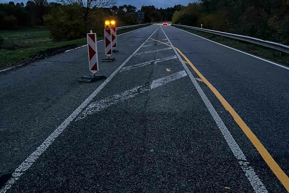 Nächtliche Straße mit Bauabsperrungen und Beleuchtung, die in die Ferne führt, mit Bäumen auf beiden Seiten, inmitten andauernder Kranarbeiten.