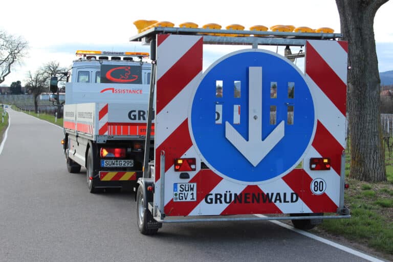 Zwei Einsatzfahrzeuge am Straßenrand mit Wegweisern, die geradeaus fahren, Teil eines Pannendienstes. Im Hintergrund Bäume und Felder.