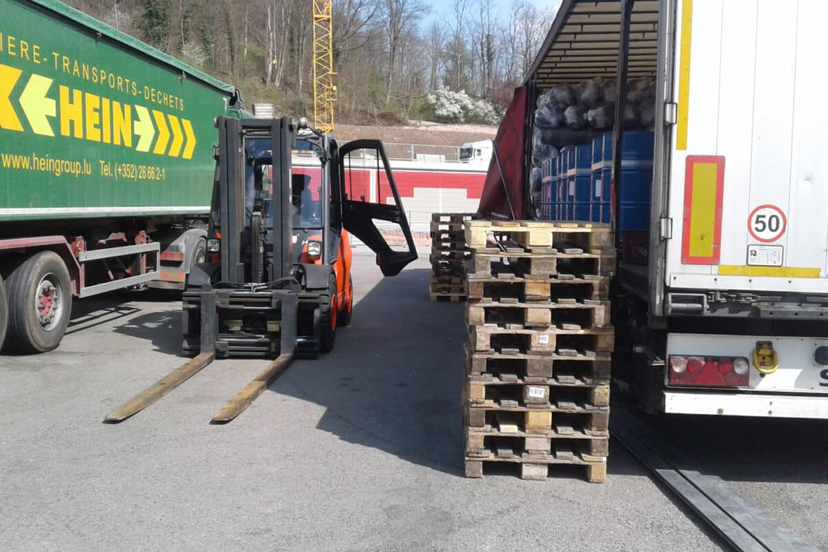 Ein Gabelstapler steht zwischen einem LKW und einem Stapel Holzpaletten in einem Industriegebiet und ist bereit für den Fahrzeugtransport.