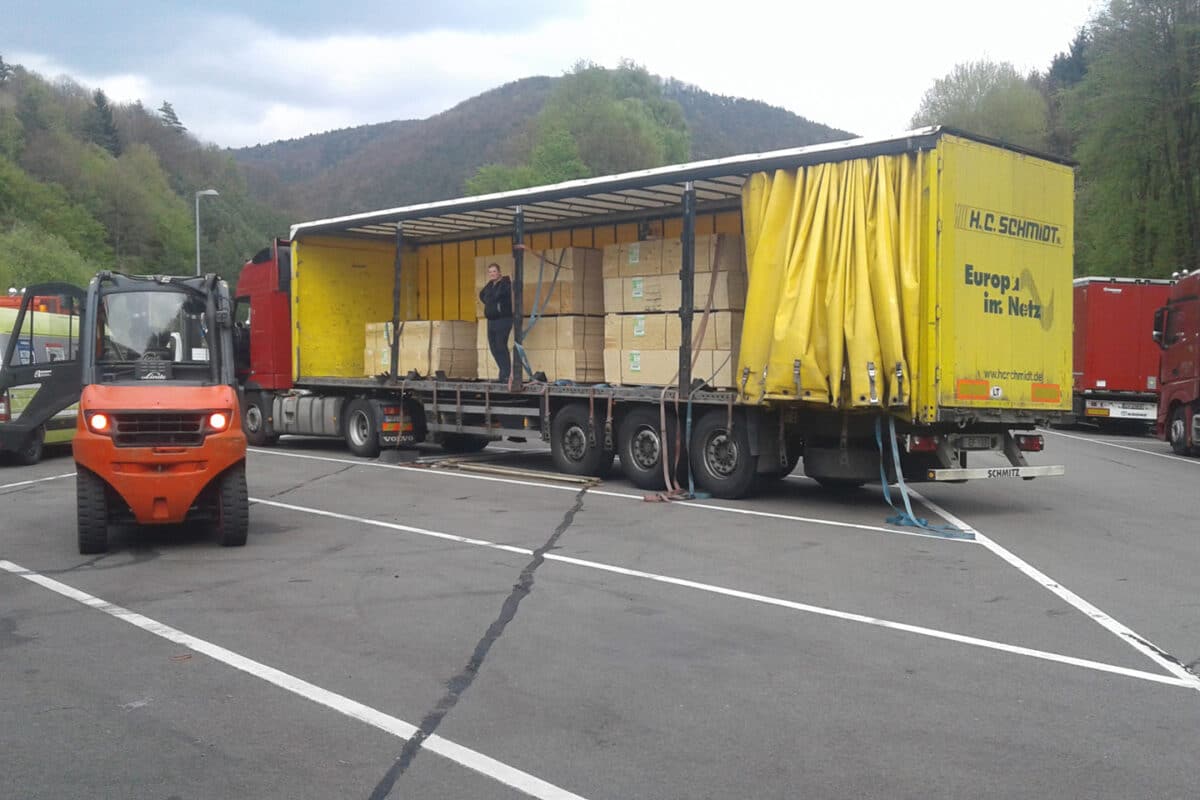 Auf einem von Bäumen und Hügeln umgebenen Parkplatz belädt ein Gabelstapler einen LKW mit Holzkisten und sorgt so zur Verkehrssicherung für einen reibungslosen Ablauf.