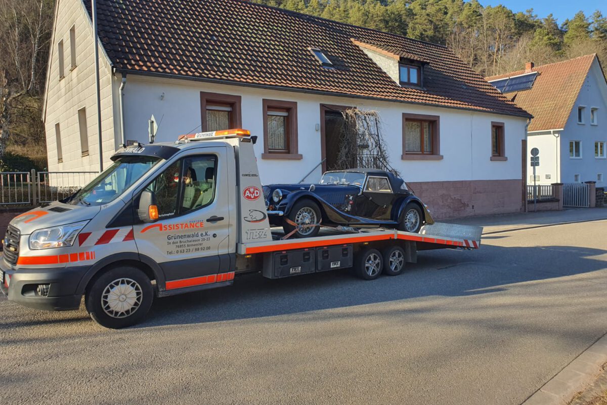 Ein Abschleppwagen mit Pritsche transportiert an einem sonnigen Tag einen Oldtimer vor Wohnhäusern und zeigt die Kompetenz unseres Bergungsdienstes.