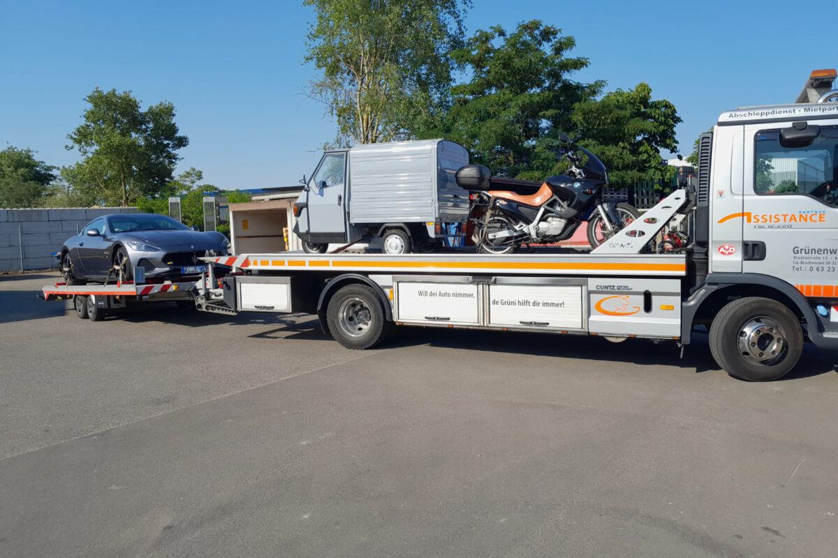 Ein Abschleppwagen von Pannendienst, der ein Motorrad und einen kleinen Lieferwagen transportiert und gleichzeitig ein Auto abschleppt, fährt durch eine sonnige Außenlandschaft.