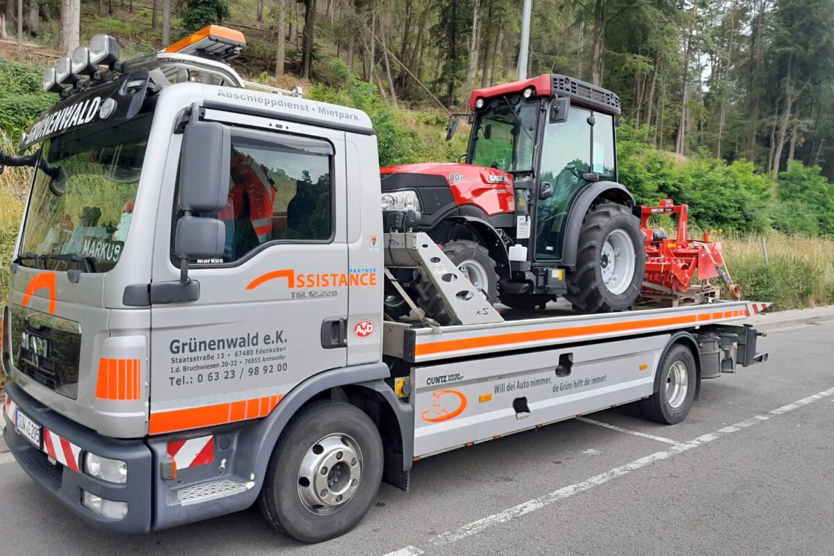 Ein Pritschen-Abschleppwagen des Abschleppdienstes fährt mit einem roten Traktor über eine von Grün umgebene Straße.