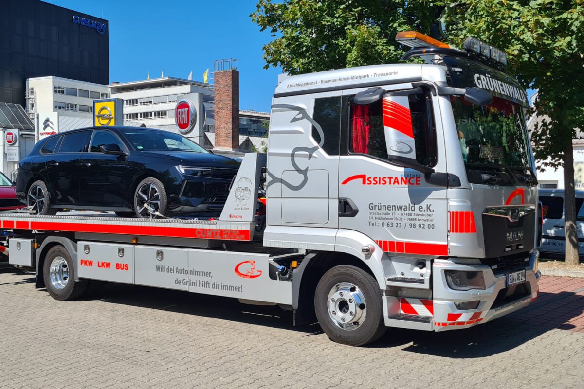 Abschleppwagen des Abschleppdienstes transportiert ein schwarzes Auto auf seiner Pritsche, an dessen Heck ein rotes Auto hängt. Städtischer Hintergrund an einem sonnigen Tag.