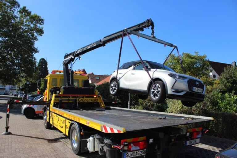 Ein weißes Auto wird von einem Abschleppkran auf eine Pritsche gehoben, um die Verkehrssicherung in der Gegend zu gewährleisten. Im Hintergrund sind Häuser und Bäume an diesem klaren Tag in Sonnenlicht getaucht.